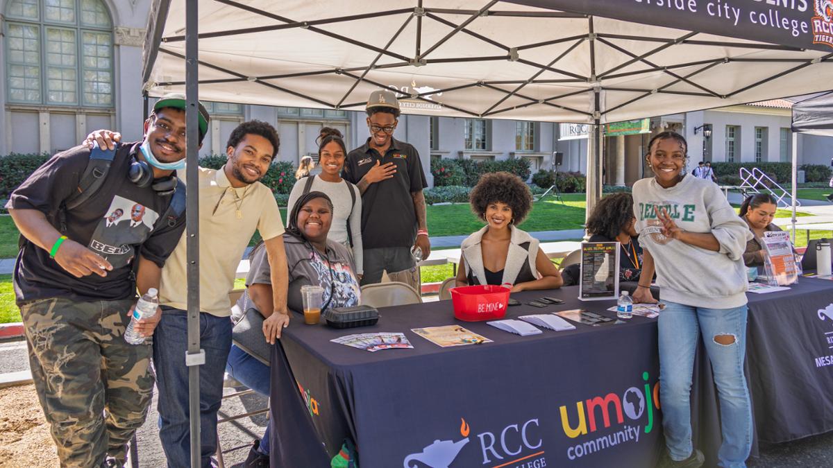 Smiling students from the Riverside City College Umoja Community posing behind a table with a purple banner displaying the RCC Umoja logo at an outdoor campus event.