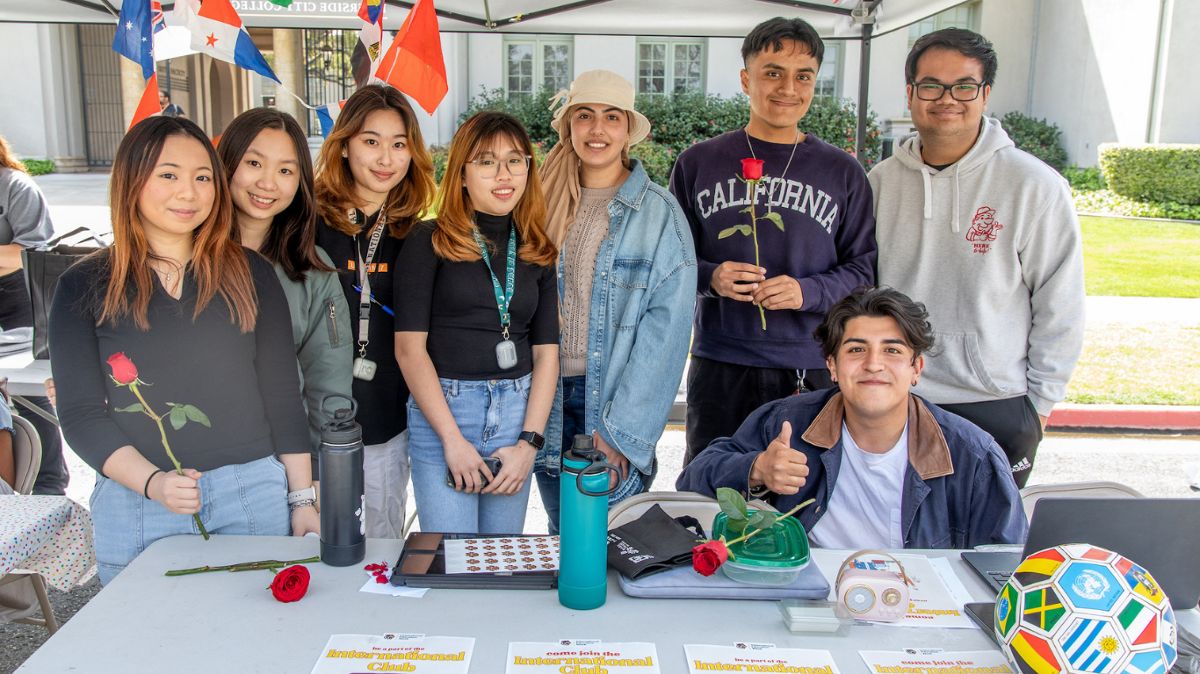 A group of students stand and sit behind a campus club table outdoors, smiling at the camera. Several hold red roses, with international flags displayed behind them and informational materials spread across the table.