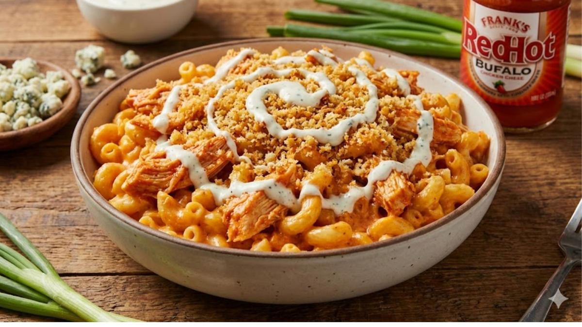 Bowl of buffalo chicken mac and cheese with elbow macaroni, shredded chicken, breadcrumb topping, and a drizzle of white sauce on a wooden table.